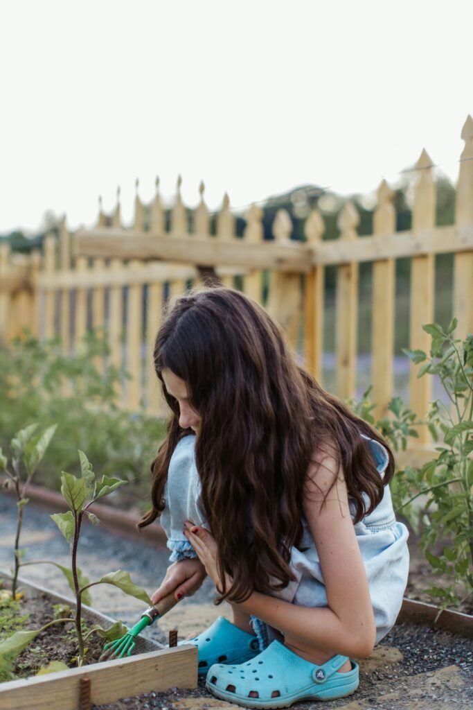 A young girl in Crocs uses a gardening fork in an outdoor garden setting.