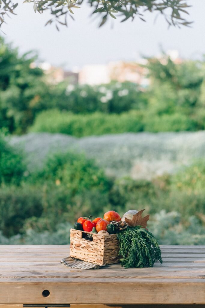A variety of organic vegetables in a woven basket on a wooden table in a lush outdoor setting.
